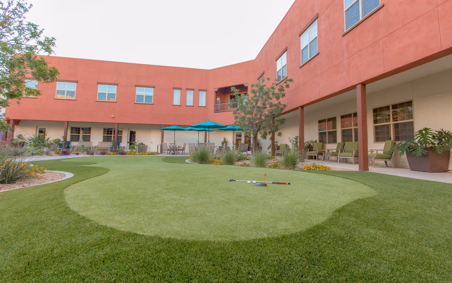Outdoor courtyard area at The Ranch Estates of Tucson featuring a putting green with golf clubs and a small flag, surrounded by green grass, plants, and flowers. The courtyard is enclosed by a two-story building with red and beige walls, windows, and a covered patio area with green cushioned chairs and tables with teal umbrellas.