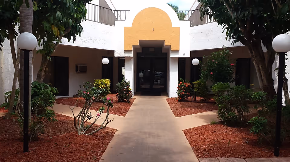 A landscaped entrance courtyard with a central walkway leading to glass doors, flanked by plants and globe lights.