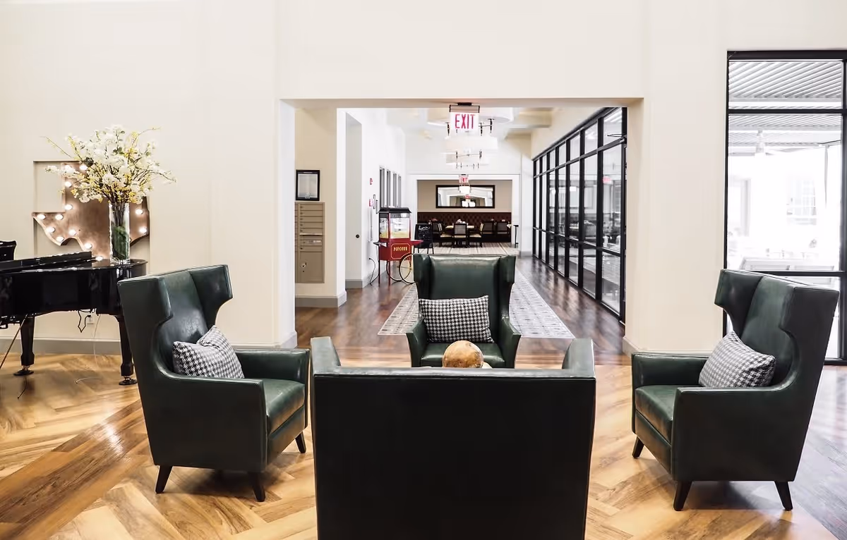 A modern and elegant seating area with four dark green leather armchairs arranged around a small table with a decorative object. The room has wooden herringbone flooring, a black grand piano with a vase of white flowers on top, and a large Texas-shaped wall decoration with lights. In the background, there is a hallway leading to a dining area with tables and chairs, and large glass windows on the right side.