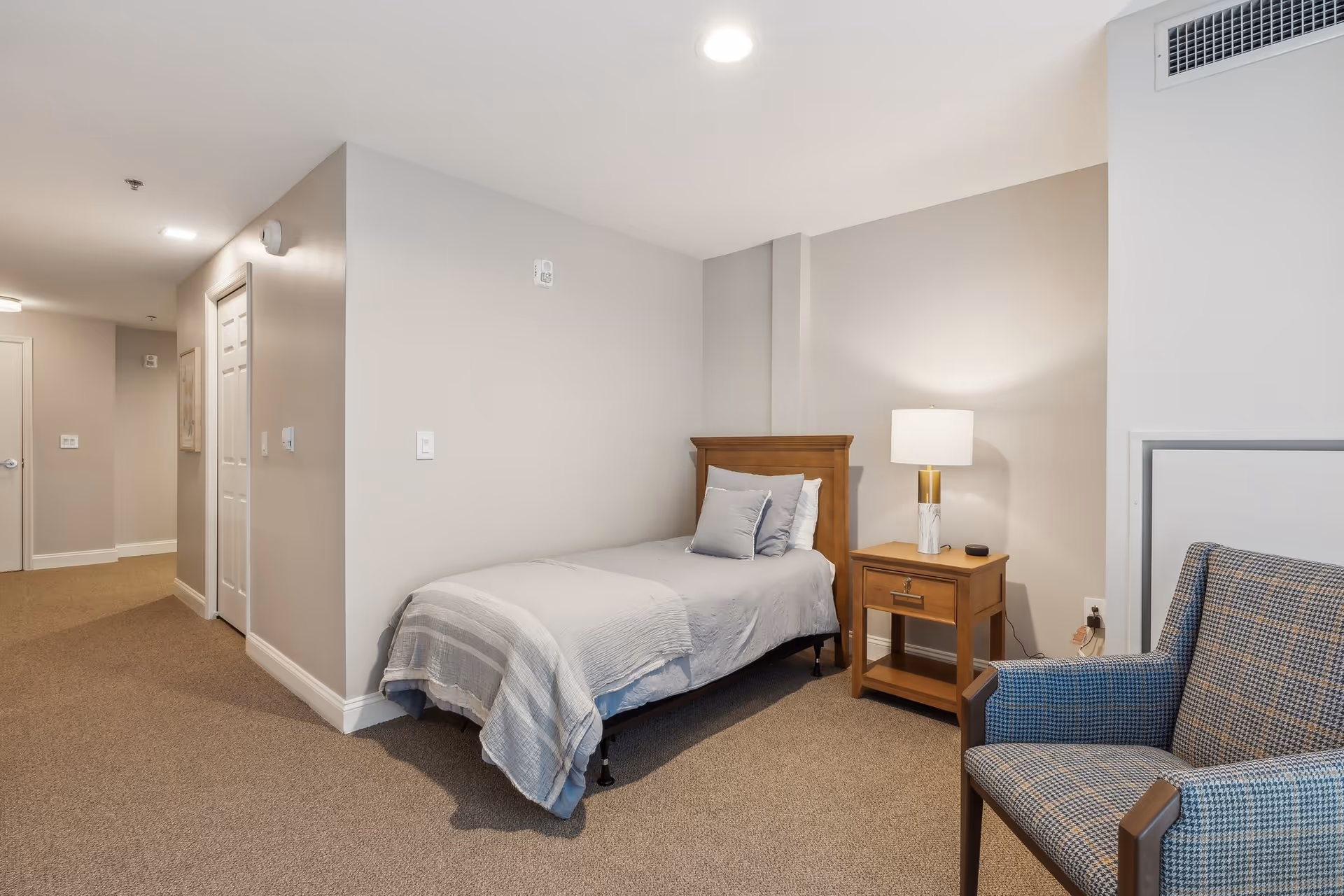 Neutral single-occupancy bedroom with a twin bed, wooden nightstand and lamp, and a patterned armchair.