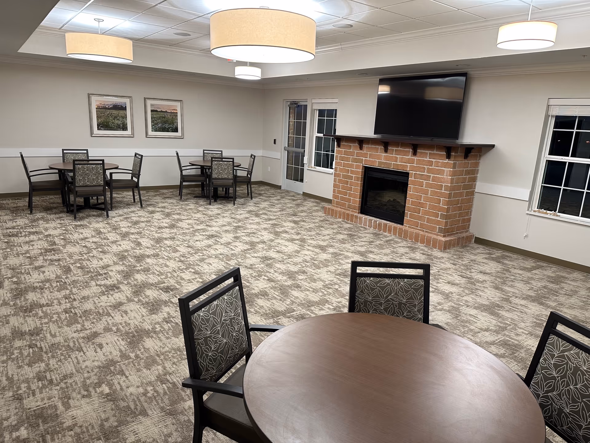A spacious common room with patterned carpet flooring, several round tables with chairs, a brick fireplace with a mounted flat-screen TV above it, two windows, and two framed landscape pictures on the wall. The room is well-lit with ceiling lights and has a neutral color scheme.