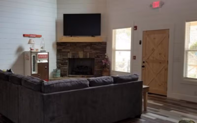 Interior view of a living room in a senior living facility with a large dark gray sectional sofa facing a stone fireplace with a mounted flat-screen TV above it. To the left of the fireplace is a vintage Coca-Cola vending machine and a small lamp. The room has white shiplap walls, two windows letting in natural light, and a wooden door with a diagonal cross design. The floor is covered with wood-patterned vinyl or laminate.