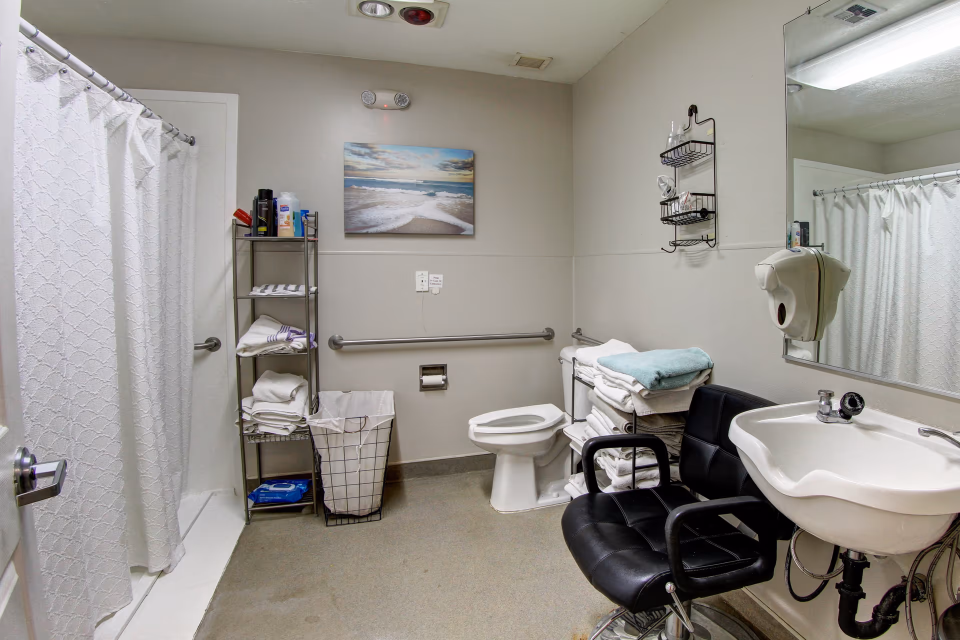 Accessible bathroom with a shower curtain, toilet and sink, grab bars, shelving stocked with towels, and a black chair.