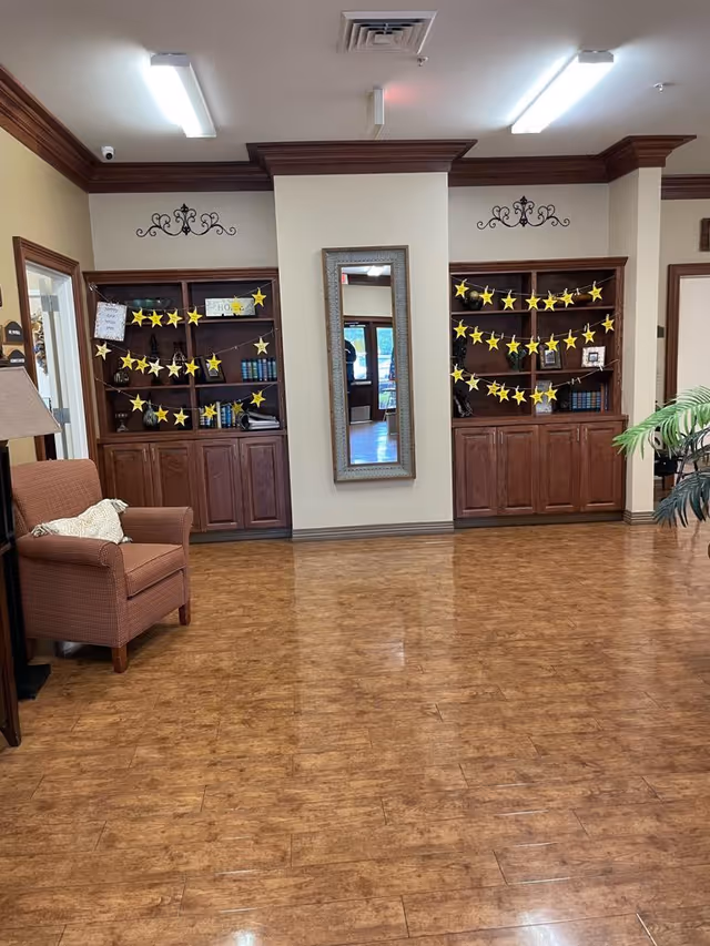 Interior view of a common area in a senior living facility with wooden flooring, two built-in wooden bookshelves decorated with yellow star garlands, a tall rectangular mirror mounted on the wall between the bookshelves, a brown upholstered armchair with a cushion on the left, and a potted plant partially visible on the right.