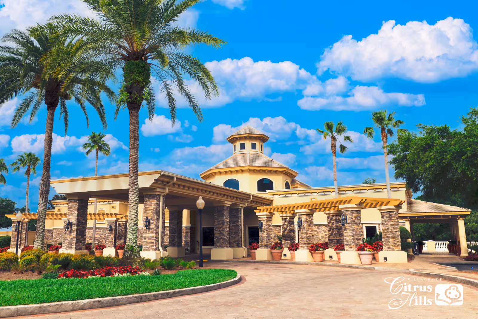 Exterior view of a senior living facility building with stone and beige walls, a covered entrance, palm trees, and landscaped greenery under a blue sky with scattered clouds.