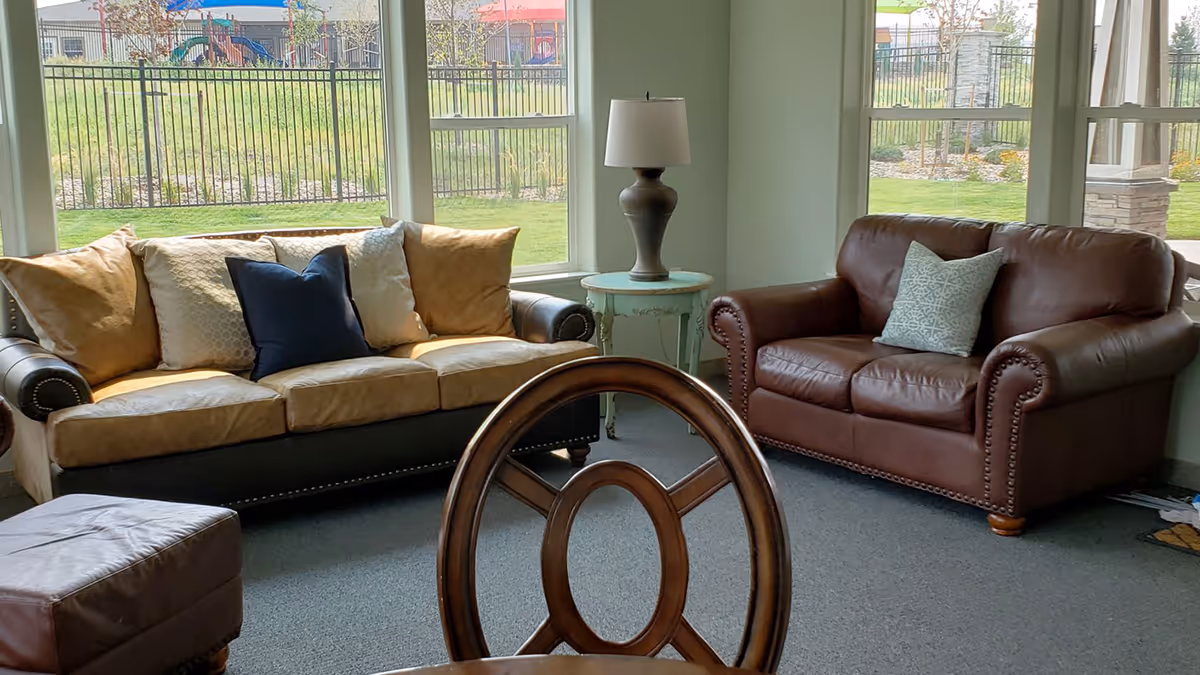 A cozy living room with a large window showing a fenced outdoor area. The room features a tan and black sofa with multiple cushions, a brown leather loveseat with a patterned cushion, a round wooden chair back in the foreground, a small round side table with a lamp, and a brown ottoman.