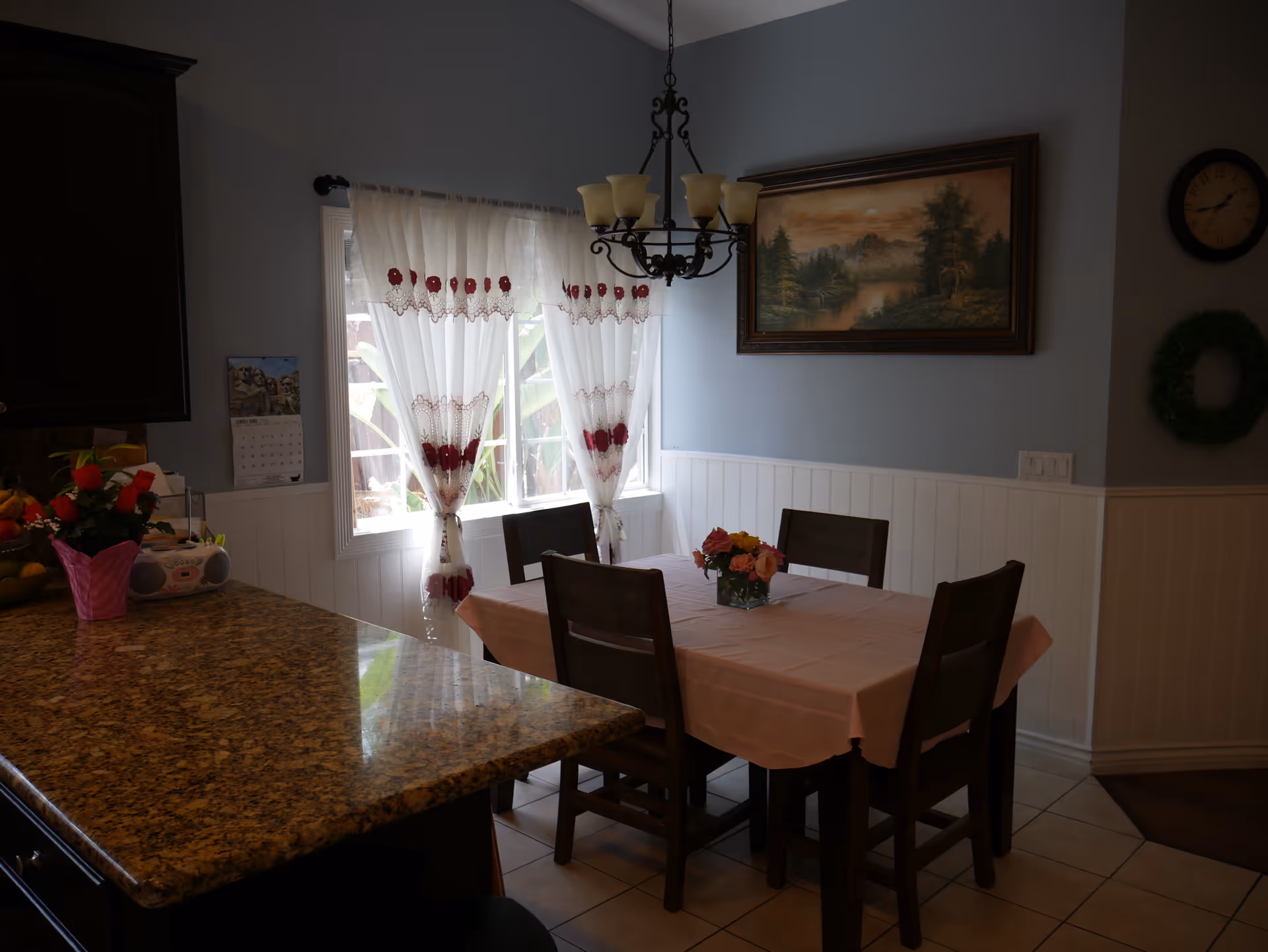 A cozy dining area with a rectangular table covered with a pink tablecloth and four wooden chairs. A small flower arrangement is placed in the center of the table. The room has light blue walls with white wainscoting, a window with sheer white curtains decorated with red accents, a hanging chandelier with five lights, a landscape painting on the wall, a clock, and a green wreath. A granite countertop with a pink flower pot and a small radio is visible in the foreground.