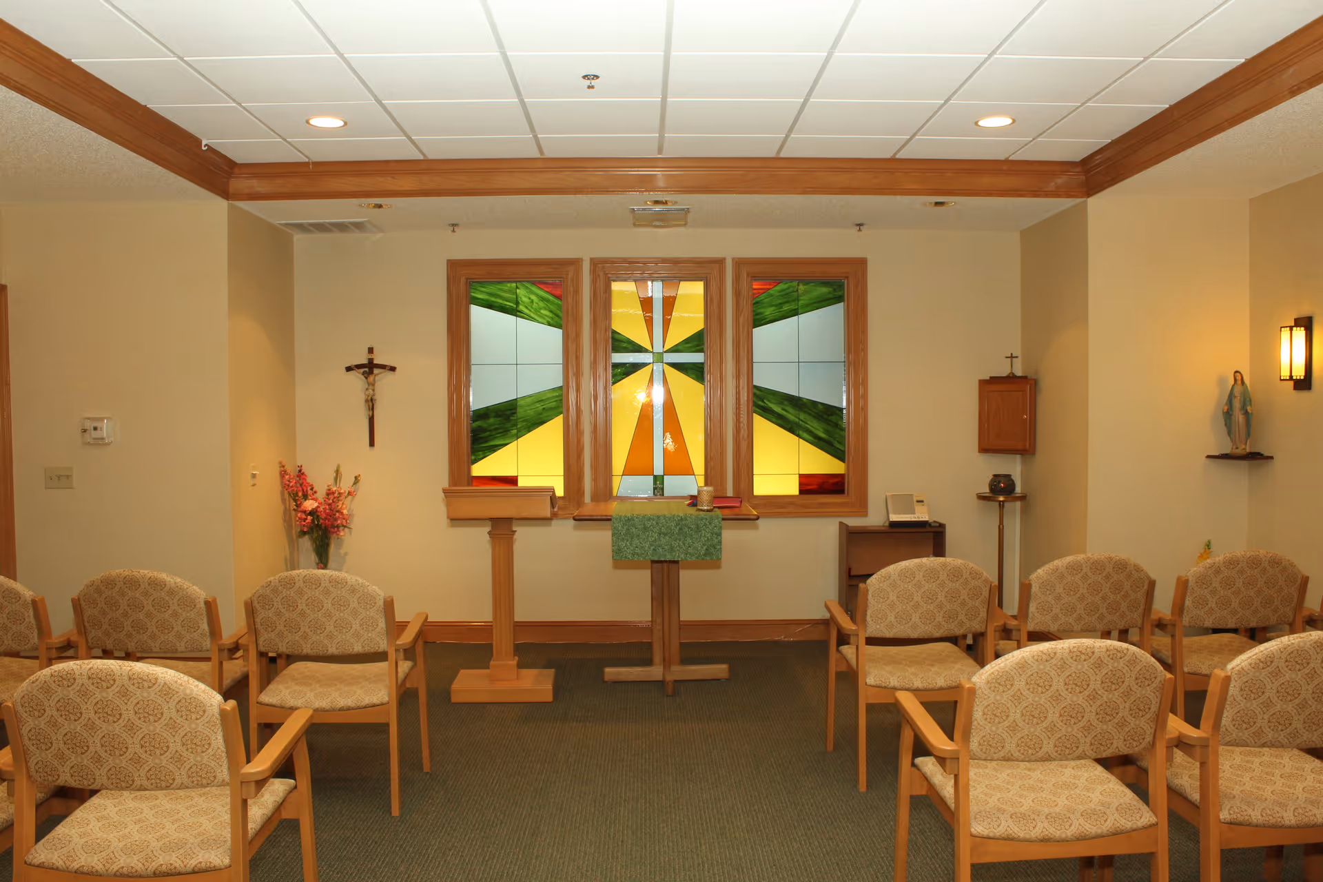 A small chapel room with rows of beige patterned chairs facing a wooden altar. Behind the altar are three stained glass windows with green, yellow, and orange geometric designs. On the left wall, there is a crucifix and a vase with flowers. On the right wall, there is a statue of the Virgin Mary on a small shelf and a wall-mounted light fixture.