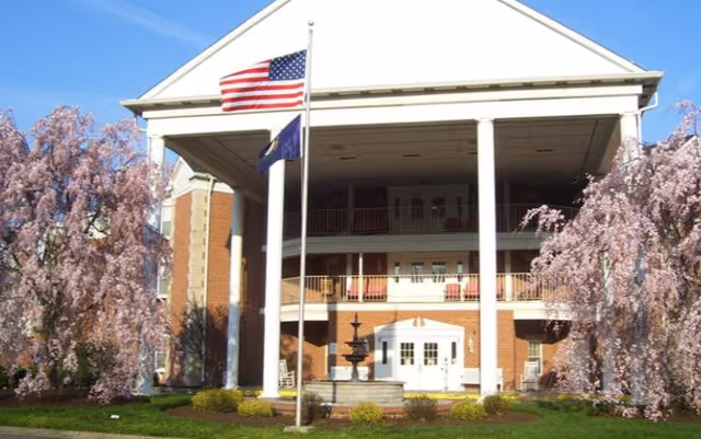 Front exterior view of Colonial Gardens facility with large white columns, two flagpoles with American and state flags, a fountain in front, and blooming pink trees on either side.