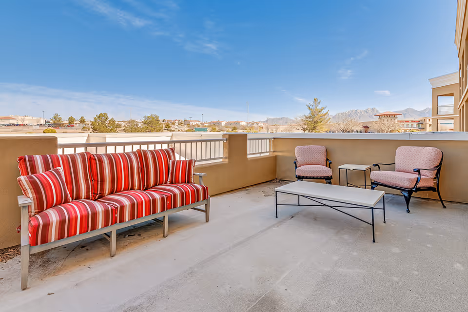 Outdoor patio area with a red and white striped cushioned bench, two pink cushioned chairs, a small side table, and a rectangular coffee table. The patio overlooks a view of trees, buildings, and distant mountains under a blue sky with some clouds.