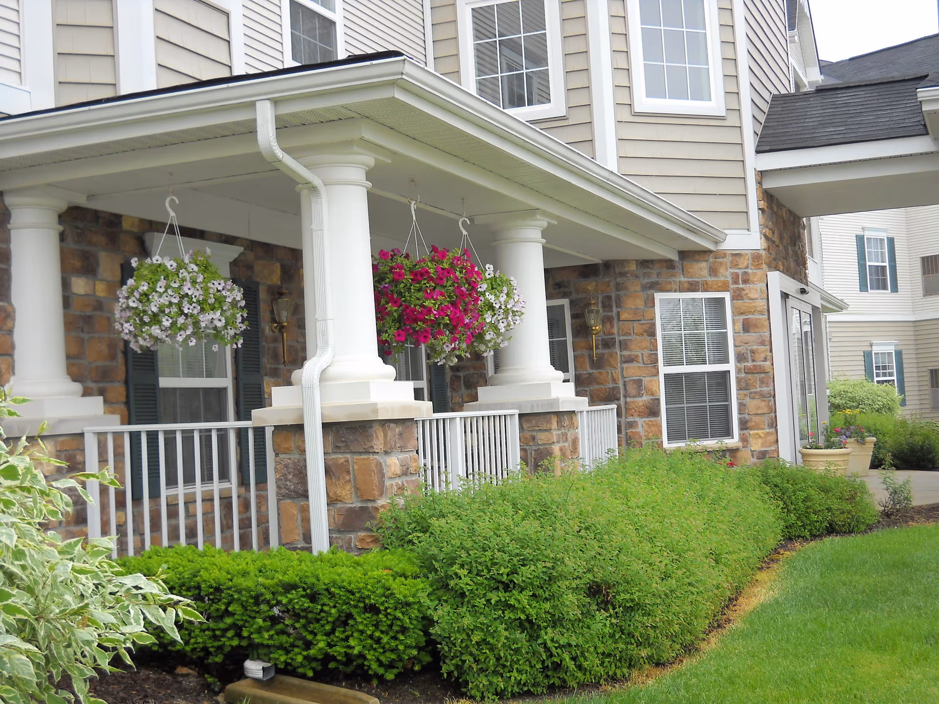 Exterior view of a senior living facility showing a porch with white columns and hanging flower baskets. The building has stone and beige siding with multiple windows. There are well-maintained green bushes and a lawn in front of the porch.