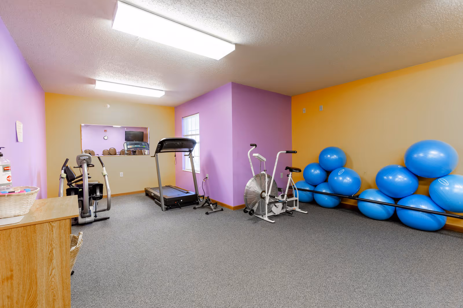 A small fitness room with exercise equipment including a treadmill, stationary bike, and elliptical machine. There are several blue exercise balls stacked against a yellow wall. The walls are painted in purple and yellow, and there is a large mirror on one wall reflecting part of the room.