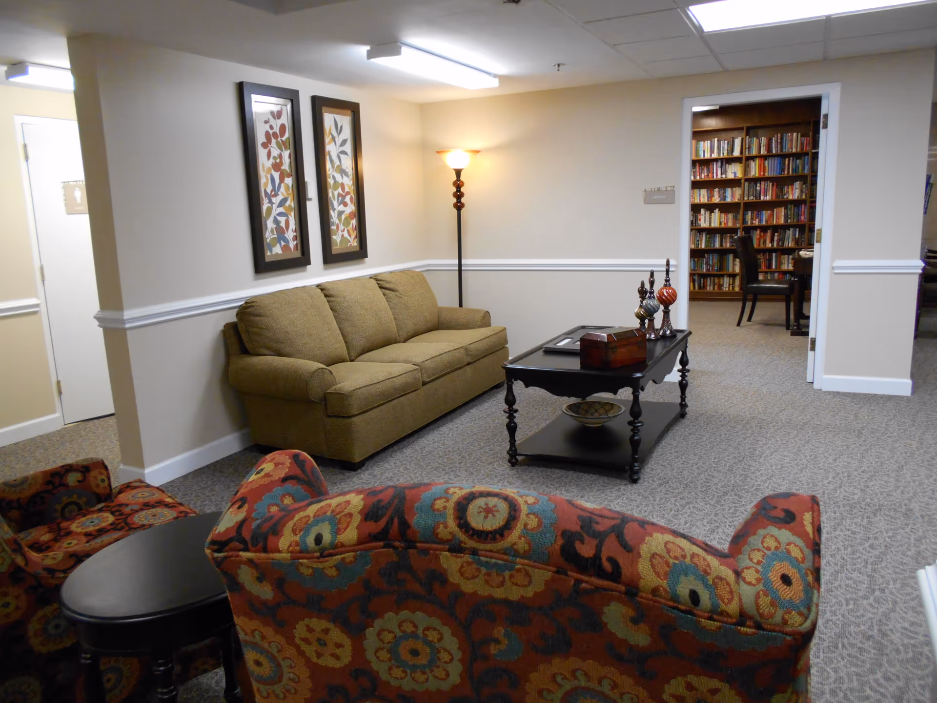 A cozy senior living common area with a beige couch, two patterned armchairs, a black coffee table with decorative items, and a floor lamp. The room has beige walls with white trim and two framed floral artworks. In the background, there is an open doorway leading to a library filled with bookshelves and a chair.