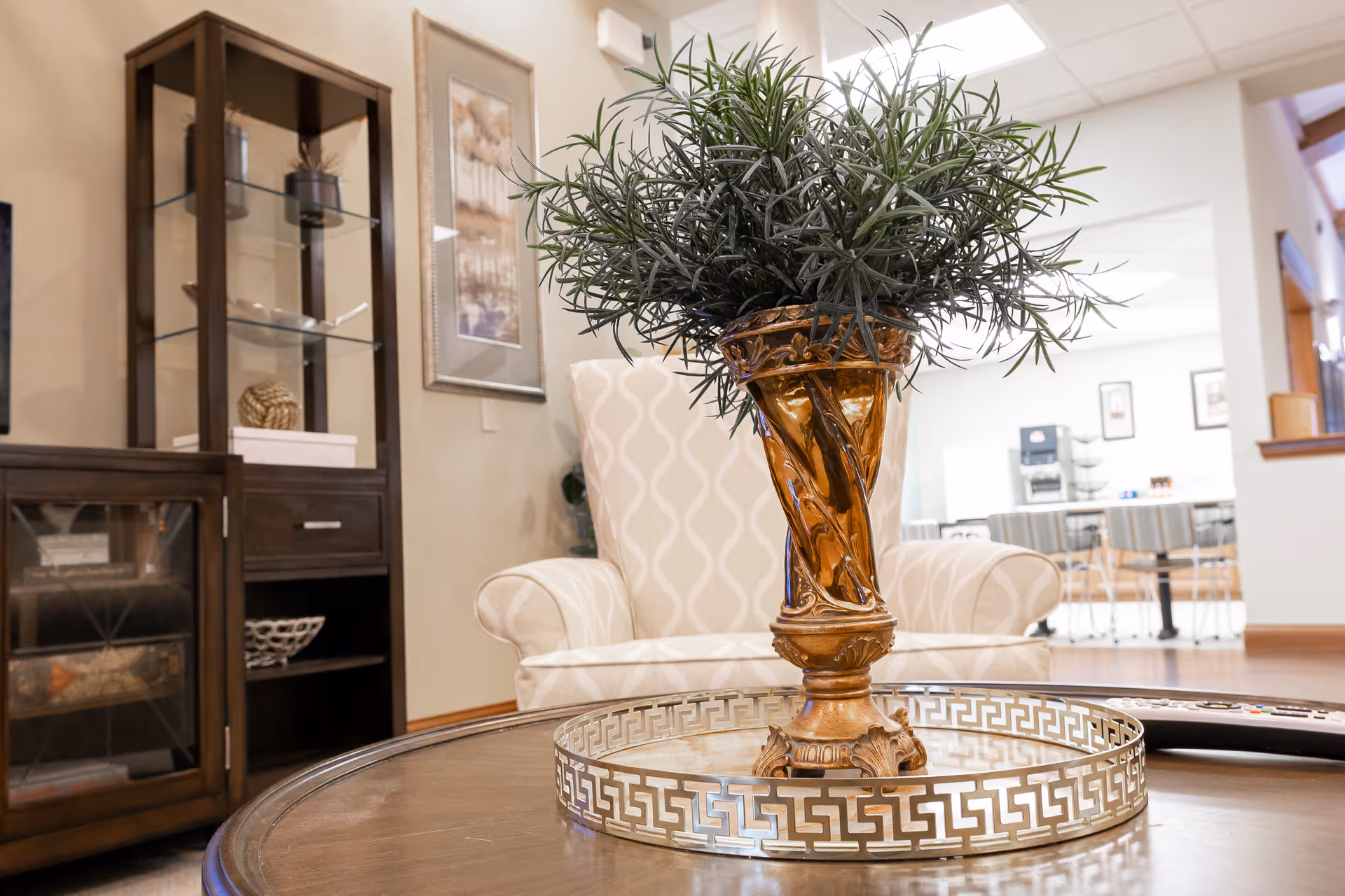 A cozy senior living room area with a decorative amber vase holding green foliage placed on a round wooden coffee table with a metallic tray. In the background, there is a beige patterned armchair, a wooden cabinet with glass shelves displaying decorative items, and a dining area with tables and chairs.