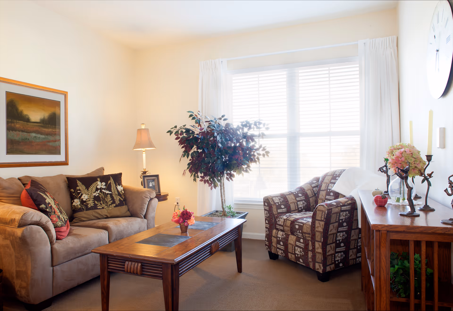 Sunlit living room with a sofa, patterned armchair, wooden coffee table, and a potted tree by a large window.
