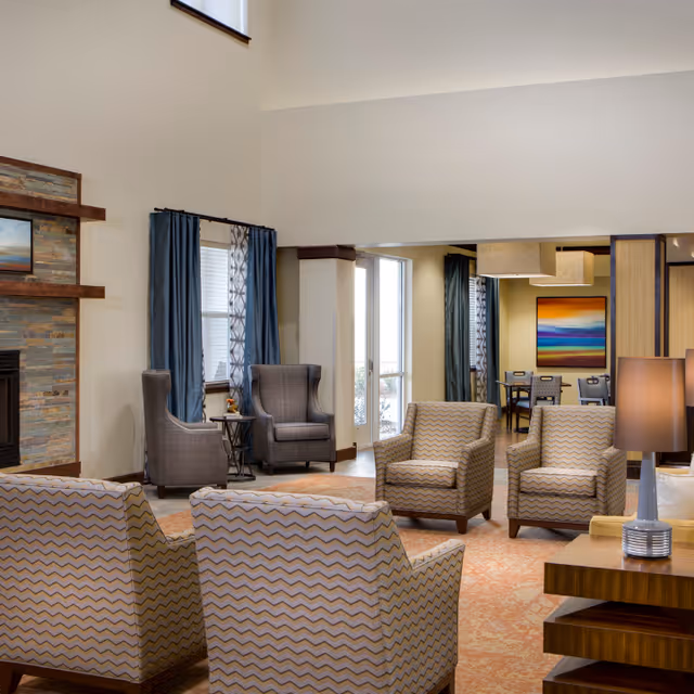 Spacious communal living room with patterned and wingback armchairs arranged near a fireplace and a doorway to a dining area.