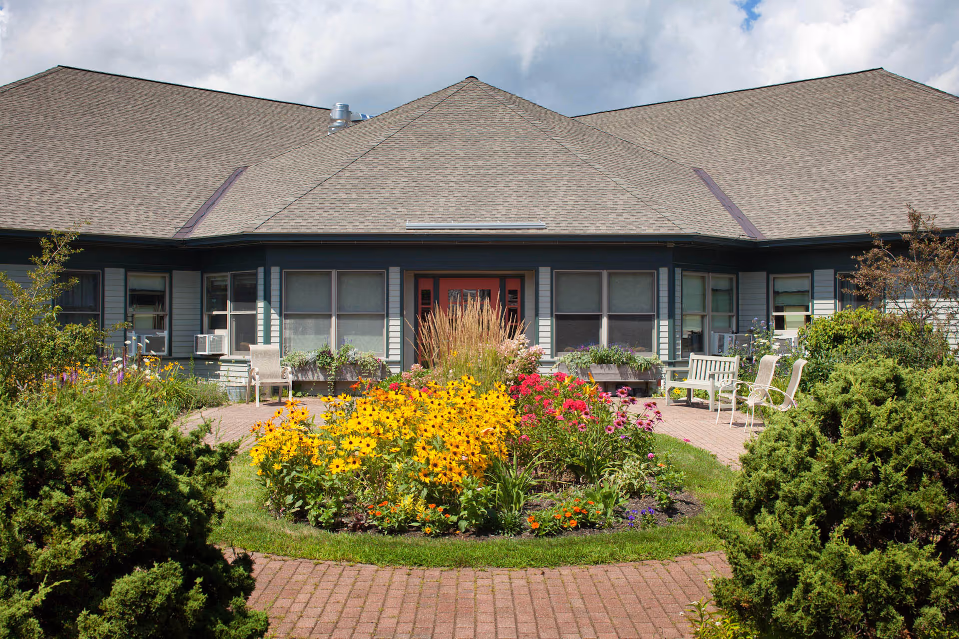 A courtyard garden with colorful flowers and green shrubs in front of a single-story building with a gray roof and multiple windows. There are several chairs and benches placed along the brick-paved walkway surrounding the garden.