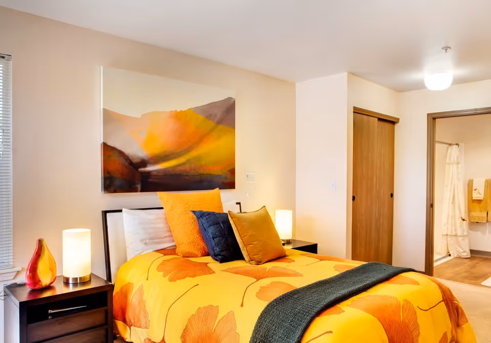 A bright bedroom with yellow-orange floral bedding, colorful throw pillows, bedside tables and lamps, a landscape painting above the bed, and an open doorway to a bathroom.