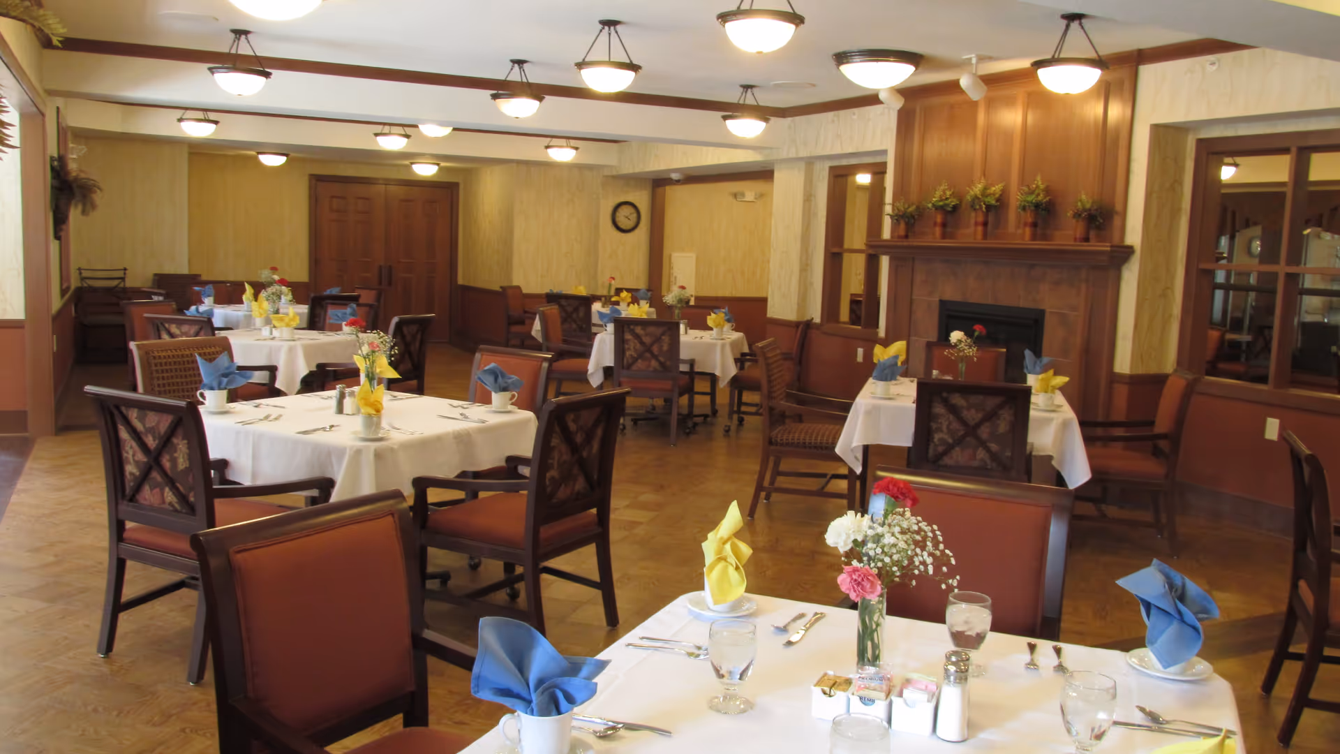 A dining room with multiple round tables covered with white tablecloths, each set with glasses, silverware, and napkins folded in blue and yellow. The room features wooden chairs, a fireplace with plants on the mantel, and warm lighting from ceiling fixtures.