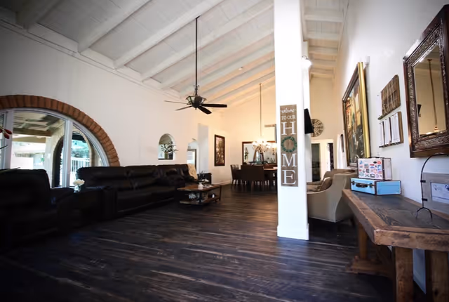 Spacious communal living room with dark wood floors, leather sofas, high beamed ceiling with ceiling fans and a dining area visible in the background.