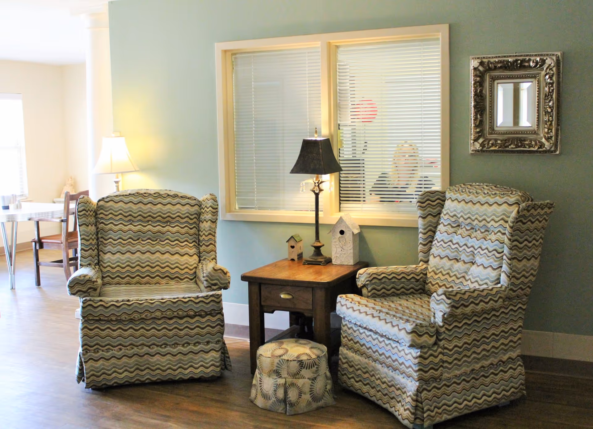 Two patterned armchairs with a small wooden side table between them, holding a lamp and two decorative birdhouses. A small patterned footstool is in front of the table. Behind the chairs is a wall with a window featuring closed blinds, through which a person is partially visible. A framed mirror hangs on the wall to the right. In the background, a dining area with a table and chairs is visible.