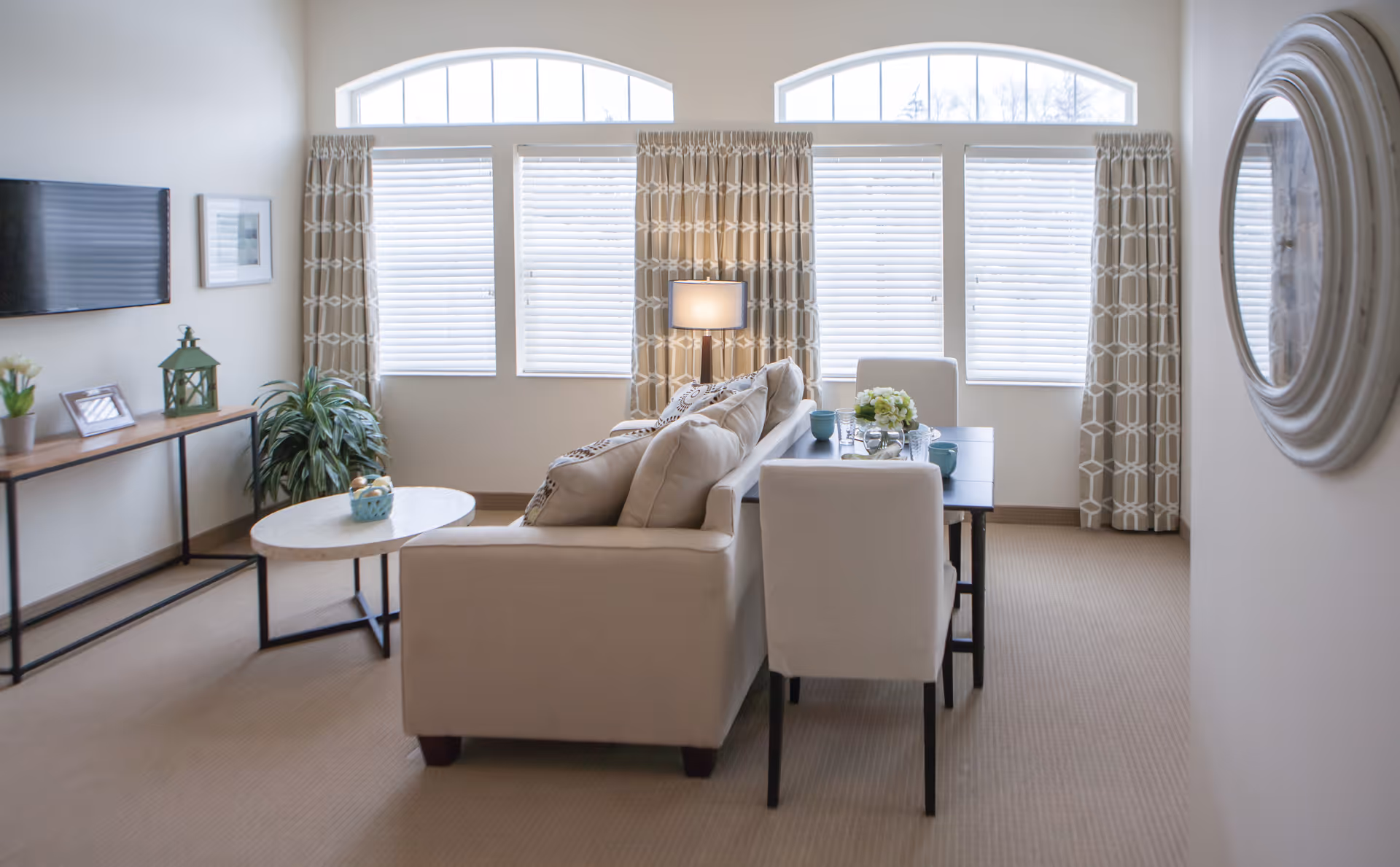 A bright living room with large windows covered by patterned curtains and white blinds. The room features a beige sofa with cushions, a small round coffee table with a decorative item, a rectangular dining table with four white chairs, a floor lamp, a wall-mounted TV, a console table with plants and picture frames, and a round decorative mirror on the wall.