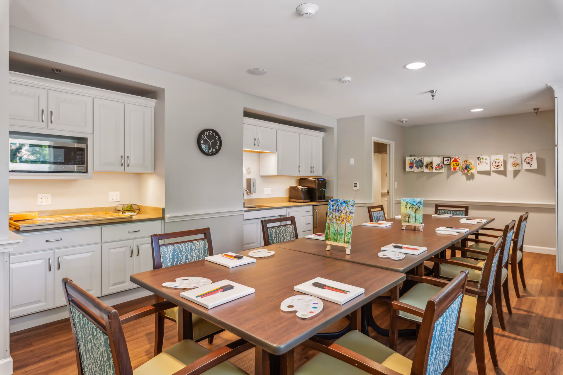 A bright dining/activity room with a long table set with art supplies and chairs, and a kitchenette with white cabinets along the wall.