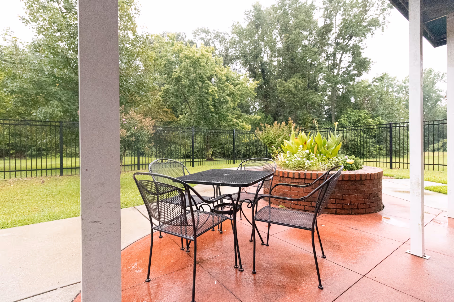 Outdoor covered patio with a metal table and chairs beside a circular brick planter and a fenced grassy yard.