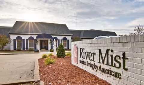 Exterior view of the River Mist assisted living facility building with a white brick sign in the foreground displaying the facility name and logo. The building has a dark roof, white walls, and a covered entrance with windows and landscaping around the driveway.