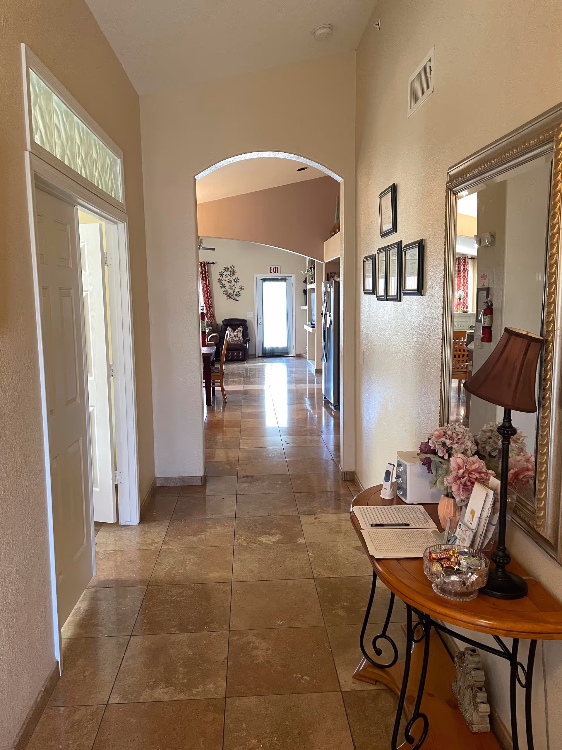 A hallway with tiled floor leading to a living area with a door at the end. On the right side of the hallway, there is a wooden table with a lamp, flowers, a bowl of candy, and some papers. Above the table is a large mirror and several framed certificates on the wall. On the left side, there is a partially open door with frosted glass above it. The living area beyond the hallway has a chair, dining table, and kitchen appliances visible.