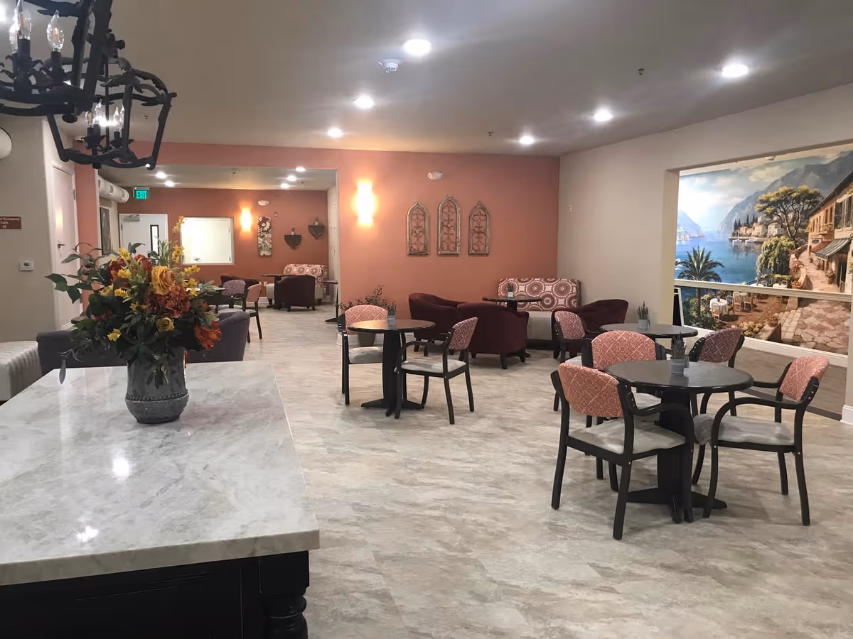A spacious senior living common area with several round tables and chairs, cushioned armchairs, and sofas. The room has a light-colored floor, peach and beige walls, and a large mural depicting a scenic coastal village. A vase with colorful flowers is placed on a marble countertop in the foreground.