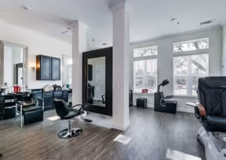 Interior view of a modern salon area with black salon chairs, large mirrors, and a bright windowed wall letting in natural light. The floor is dark wood, and the space is clean and well-lit.