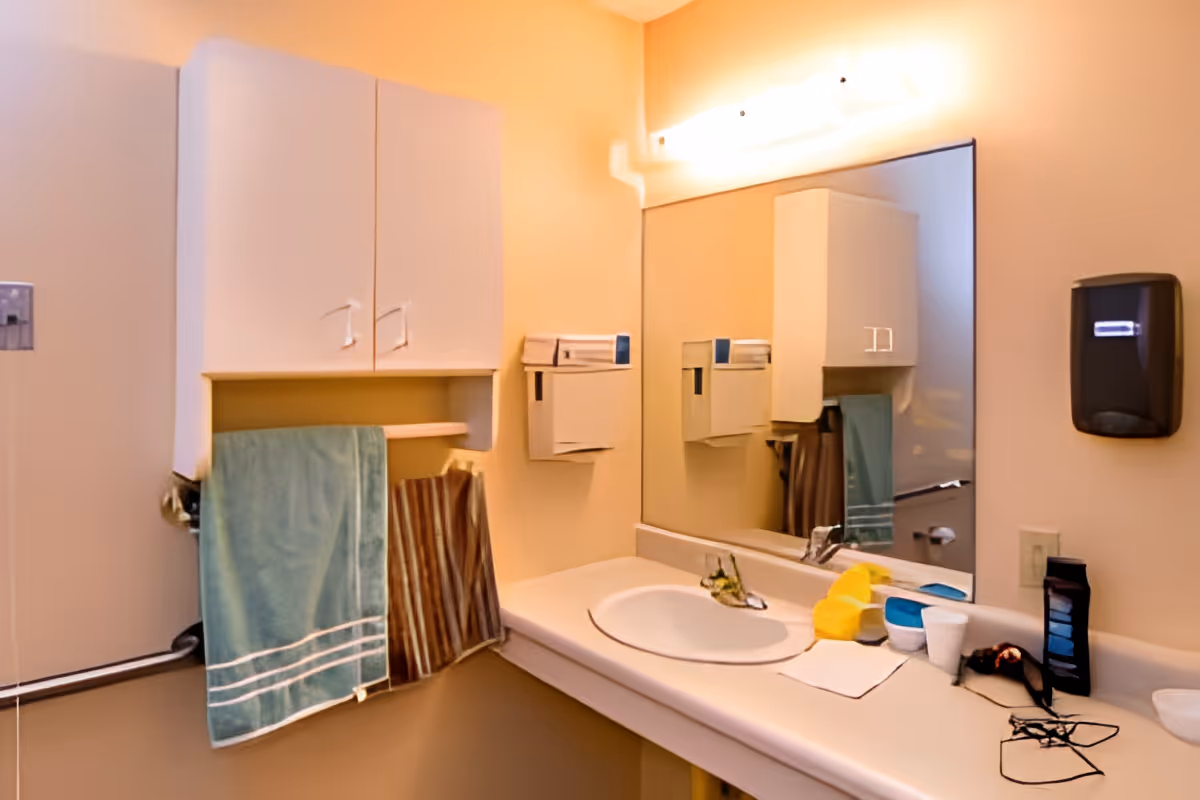 A bathroom sink area with a large mirror above the counter. There are two towels hanging on a rack below a white cabinet. On the counter, there are various personal care items, including a soap dispenser, cups, and a few small containers. The walls are painted a light beige color, and there is a wall-mounted soap dispenser on the right side.