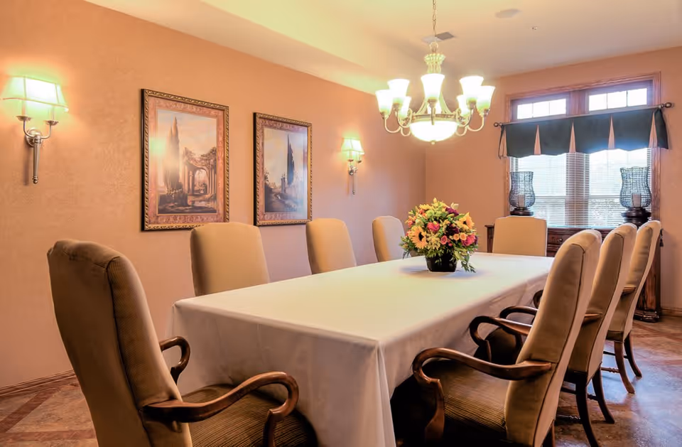 A dining room with a long rectangular table covered with a white tablecloth, surrounded by eight cushioned chairs with wooden armrests. A floral centerpiece is placed in the middle of the table. The room has warm beige walls with two framed paintings, wall sconces, a chandelier hanging above the table, and a window with a dark valance letting in natural light.