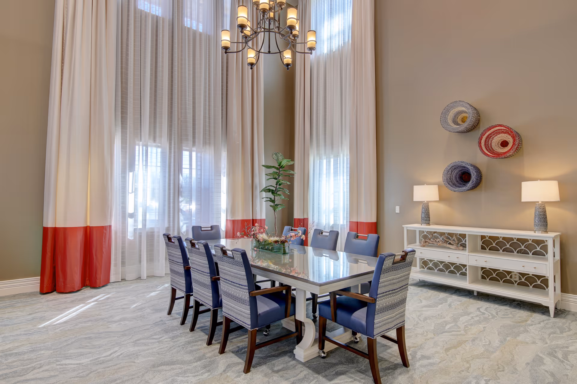 A bright dining room with a large rectangular glass table surrounded by eight blue cushioned chairs. The room features tall windows with white and red curtains, a chandelier hanging from the ceiling, a white sideboard with two lamps, and three colorful woven baskets mounted on the wall above the sideboard.
