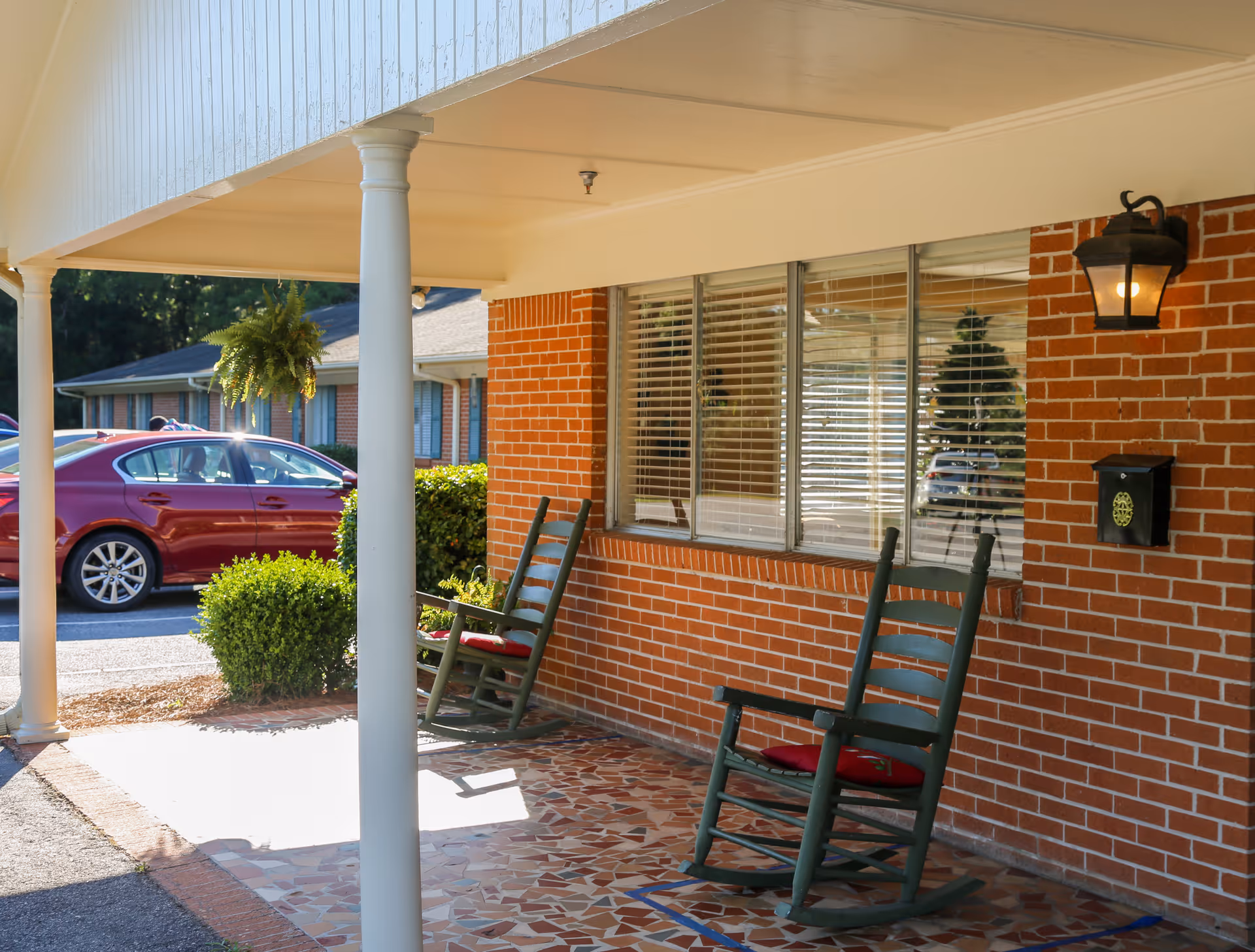 Covered porch area with two green rocking chairs with red cushions placed on a tiled floor. The porch has white pillars and a brick wall with windows and a black lantern light fixture. A red car and some greenery are visible in the background.