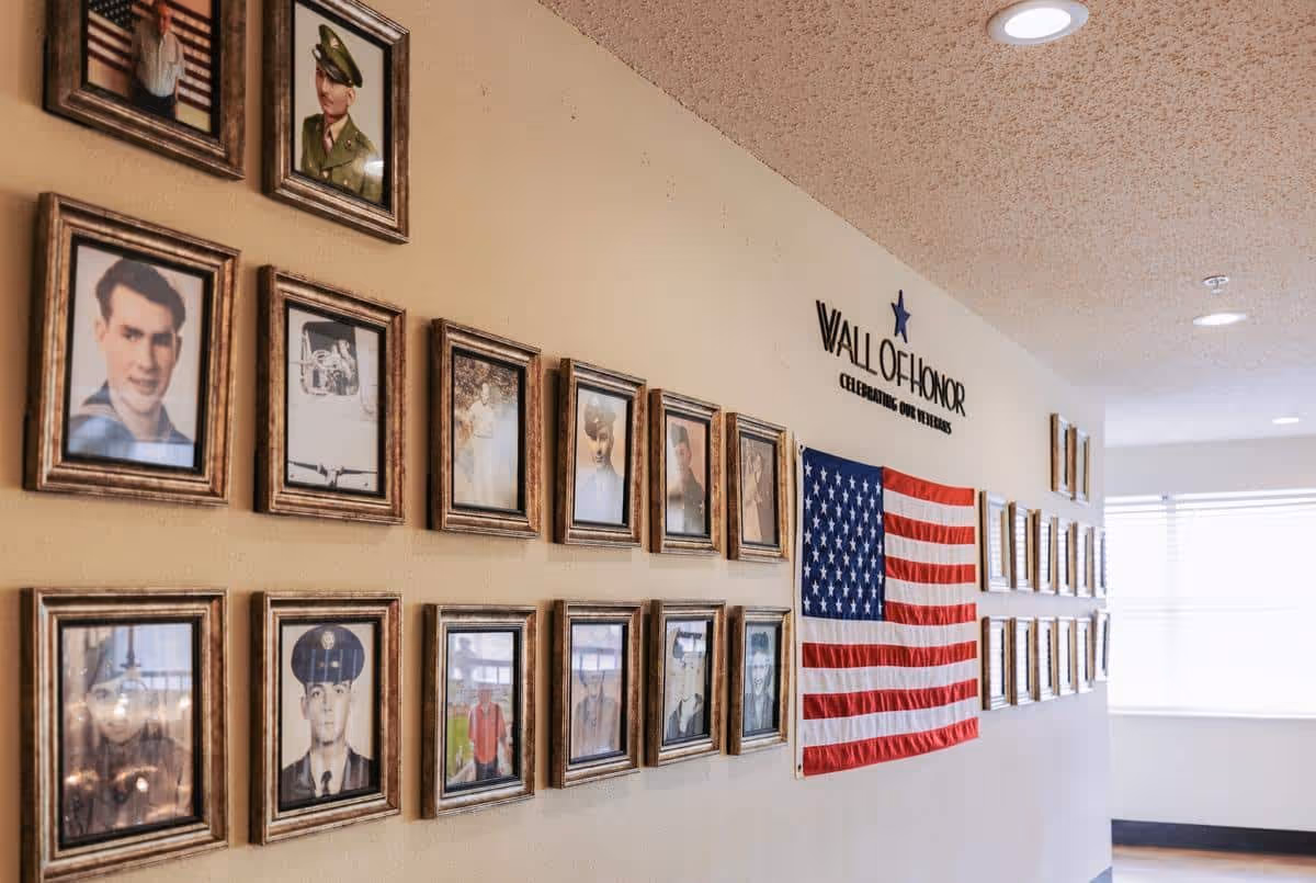 A hallway wall displaying a 'Wall of Honor' with framed photographs of veterans and an American flag. The wall honors veterans with the text 'Wall of Honor Celebrating Our Veterans' above the flag and photos.