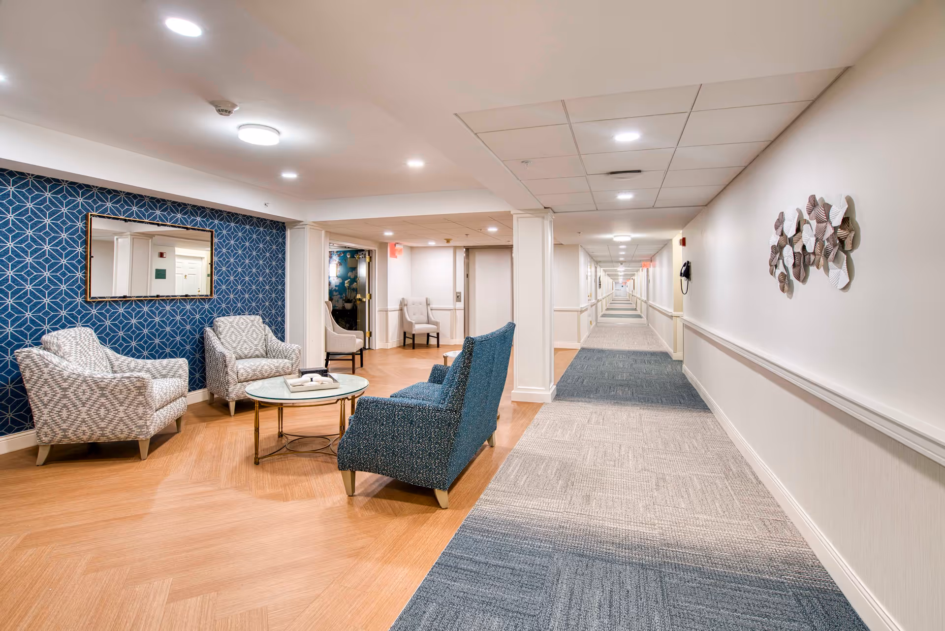 A long, well-lit hallway in a senior living facility with a seating area on the left side. The seating area includes two patterned armchairs, a blue upholstered chair, and a round glass coffee table. The wall behind the seating area has a blue geometric patterned wallpaper and a rectangular mirror. The hallway has a carpeted floor with a mix of blue and beige tones, white walls, and ceiling lights. Decorative wall art is visible on the right wall.