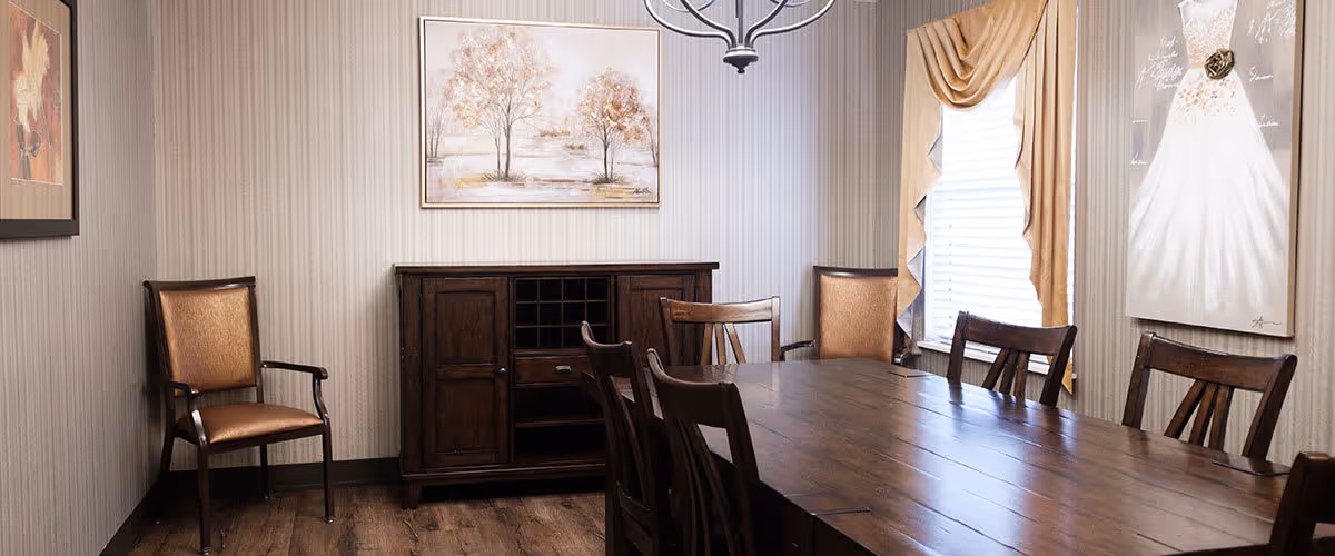 A dining room with a long wooden table surrounded by wooden chairs. The room has beige striped wallpaper, two framed paintings on the walls, and a window with beige curtains. There is a dark wooden sideboard against the wall and a chandelier hanging from the ceiling.
