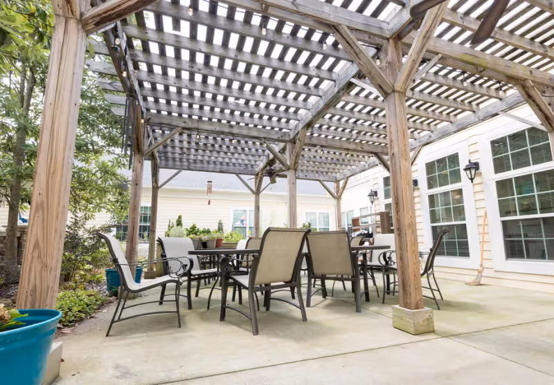 Outdoor patio area at Cadence at Wake Forest featuring a wooden pergola with lattice roof, several metal and fabric chairs around a rectangular table, potted plants, and a building with large windows in the background.