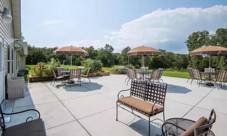 Outdoor patio with metal chairs, tables under umbrellas and a bench overlooking a grassy landscape.