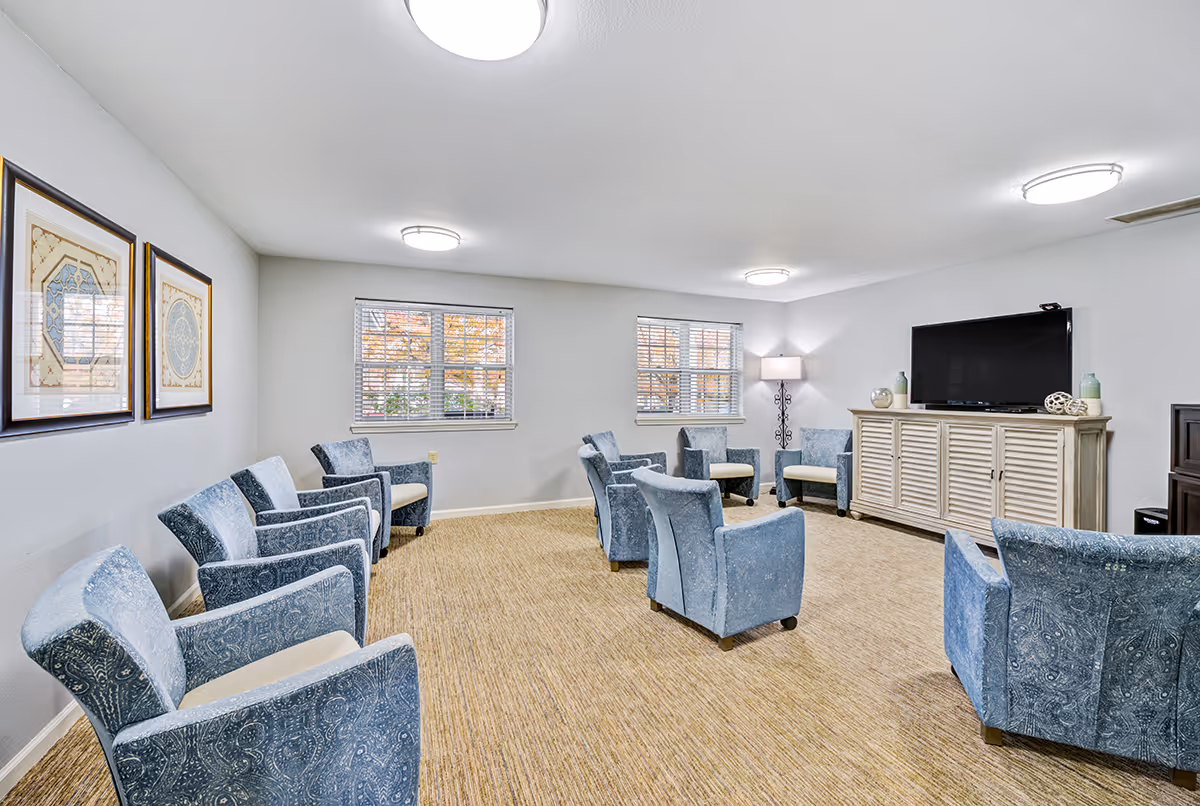 A spacious living room with multiple blue patterned armchairs arranged in a circle on a beige carpet. The room has white walls with two framed artworks, two windows with blinds, a floor lamp, and a large flat-screen TV on a light-colored cabinet.