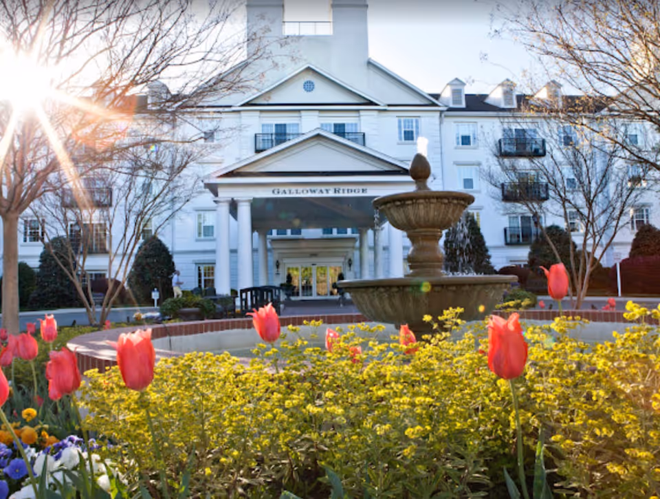 Front exterior view of Galloway Ridge at Fearrington senior living facility with a water fountain in the foreground surrounded by blooming red tulips and yellow flowers, trees without leaves, and sunlight shining from the left.