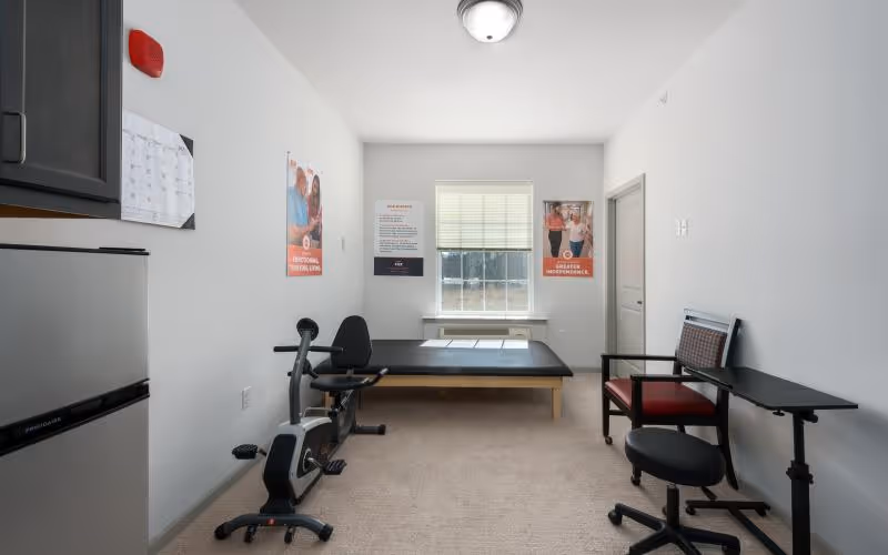 A small therapy room with a stationary exercise bike, a padded therapy table, a chair with an attached table, and a stool. The room has white walls with three posters, a window with blinds, and a small refrigerator in the corner.