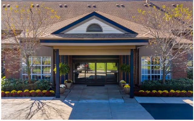Front entrance of a brick building with a covered portico, glass double doors, potted plants and landscaped flowerbeds.