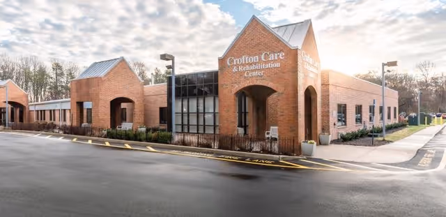 Front exterior of a brick senior care and rehabilitation building with arched entrance, windows and an empty driveway under a partly cloudy sky.