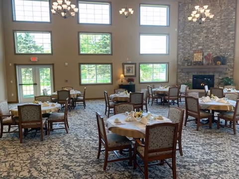 A spacious dining room with multiple round tables covered with white tablecloths and set with cups, plates, and napkins. The room features large windows allowing natural light to fill the space, a stone fireplace on the right wall, and elegant chandeliers hanging from the high ceiling. The carpet has a floral pattern, and the walls are painted in a warm beige tone.
