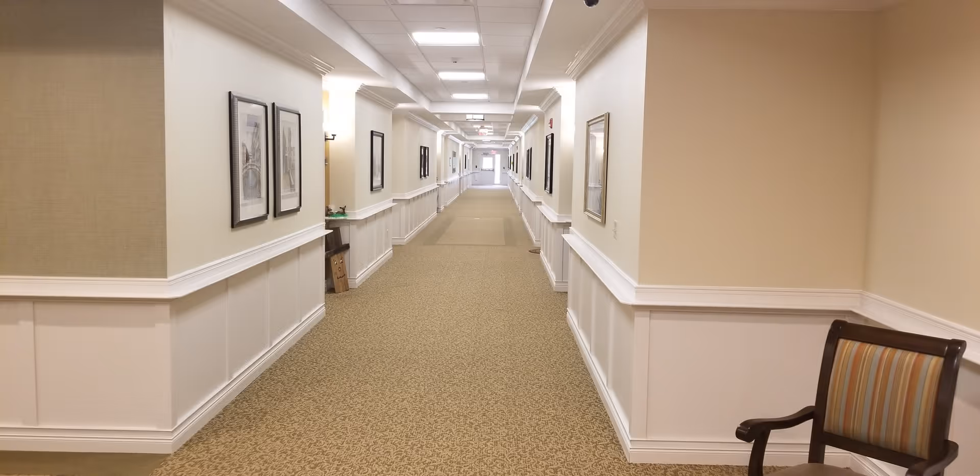 A long, well-lit hallway in a senior living facility with beige walls, white wainscoting, and patterned carpet. Several framed pictures hang on the walls, and a striped upholstered chair is positioned on the right side near the foreground.