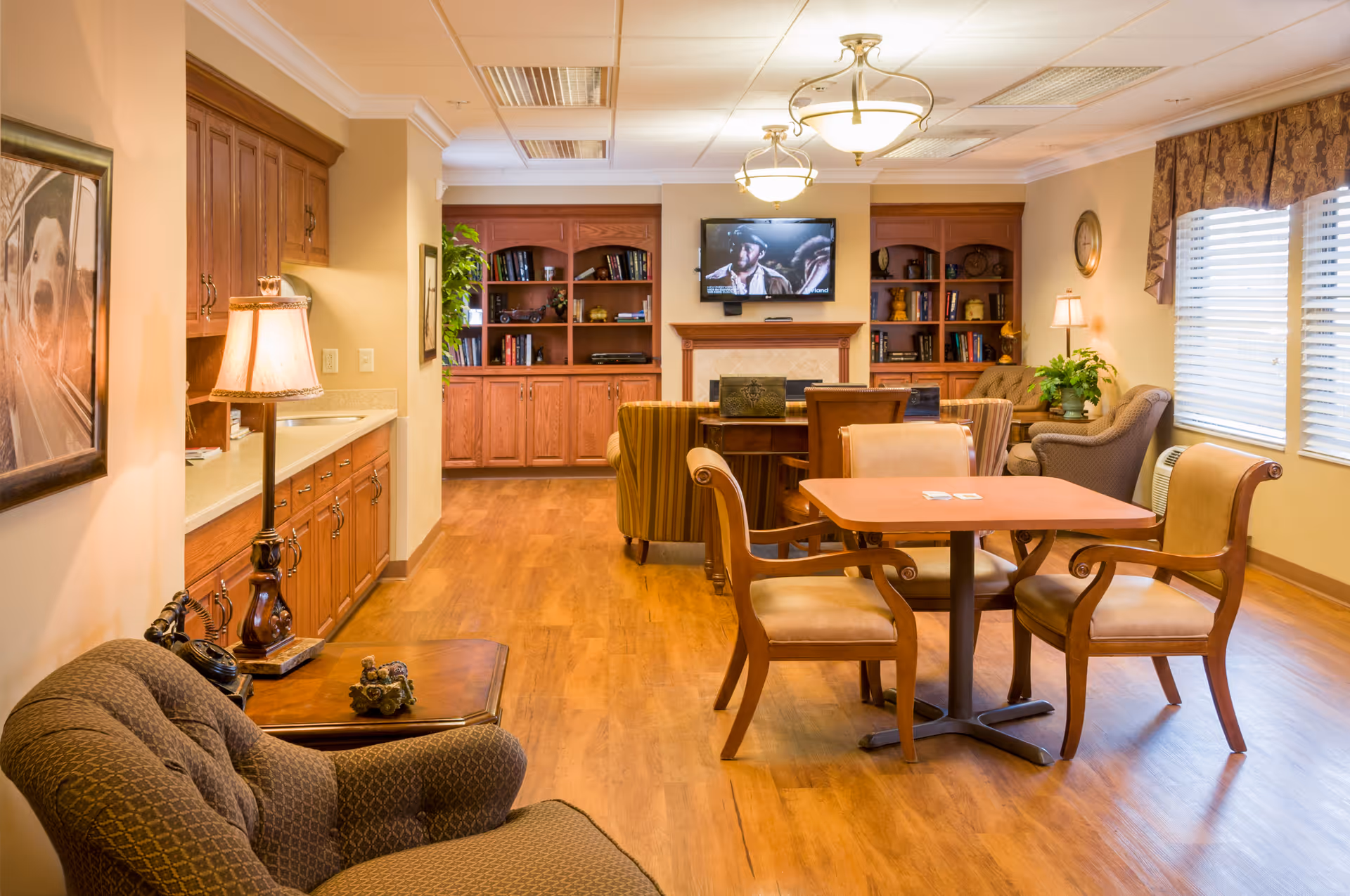 A cozy living room area in a senior living facility with wooden flooring, a table with four chairs, a comfortable armchair, a side table with a lamp, built-in wooden bookshelves, a wall-mounted TV above a fireplace, and large windows with blinds and valances letting in natural light.