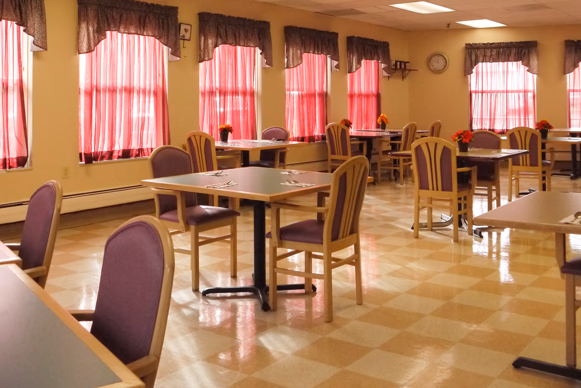 A dining room with multiple tables and chairs arranged neatly. The chairs have purple cushions and wooden frames. The room has several windows with red curtains allowing natural light to enter. Small flower arrangements are placed on some tables, and a clock is visible on the wall.